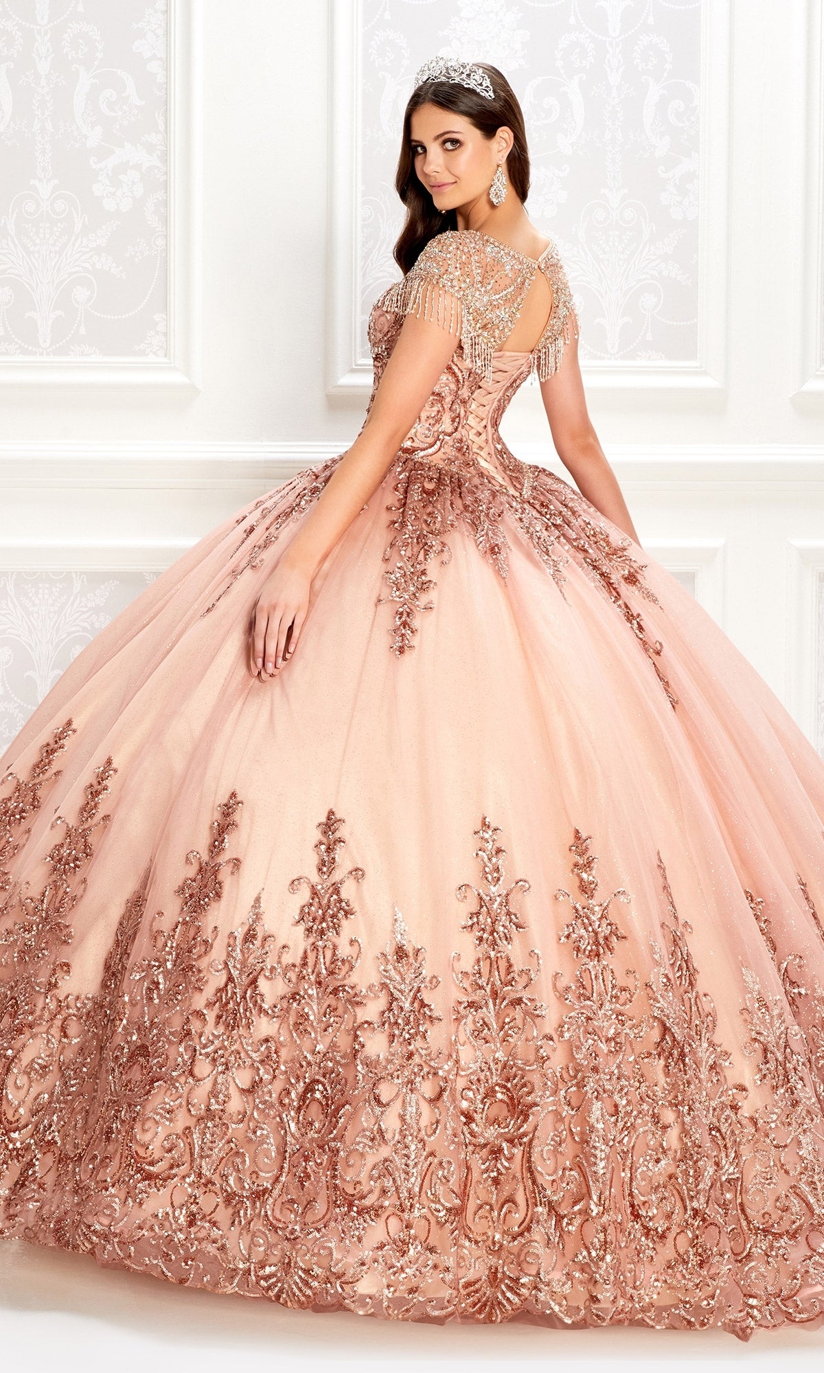 A young woman in the Princesa PR22026 short-sleeve quinceanera dress with a beaded bodice and rose gold embroidery stands in an elegant white room, wearing a tiara and glancing over her shoulder.